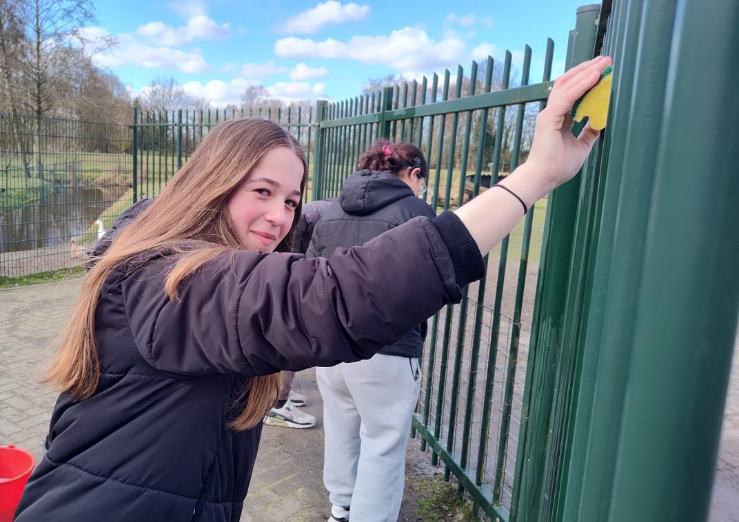 Twee personen reinigen een groen metalen hekwerk buiten, met een rode emmer op de grond.