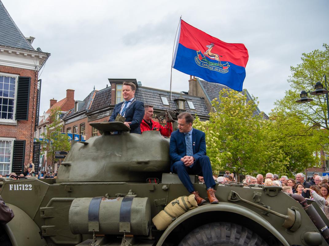 Historisch militair voertuig met mensen erop tijdens een herdenkingsevenement, met een rood-blauw vlag en omringd door toeschouwers in een stadsomgeving