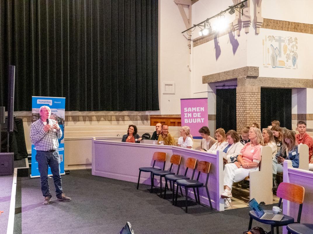 Spreker met microfoon op podium voor publiek; banners met 'Noardeast Ferbûn' en 'SAMEN IN DE BUURT' op de achtergrond.
