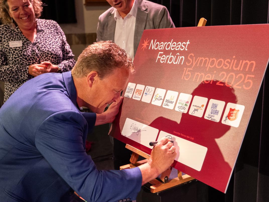 Persoon in blauw pak tekent op bord met 'Noardeast Ferbûn Symposium 15 mei 2025', terwijl twee anderen toekijken