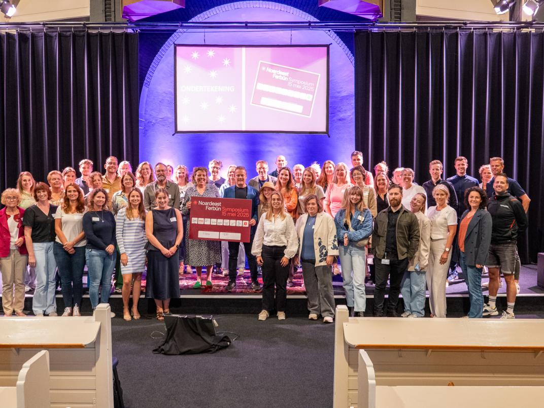 Grote groep mensen op podium voor scherm met 'Nederlands voor anderstaligen'; centrale persoon houdt een bord vast.