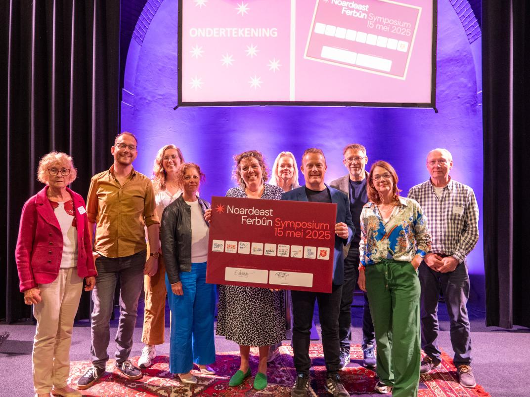 Groep mensen poseert op een podium bij het Noardeast Ferbûn Symposium op 15 mei 2025, met een groot bord en projectiescherm op de achtergrond