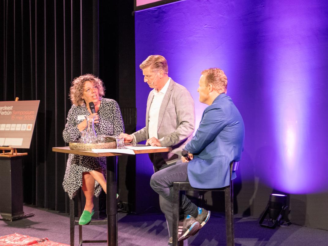 Wethouder Lysbeth Vellinga spreekt in een paneldiscussie op een podium, zittend aan een hoge tafel met twee andere personen. Ze houdt een microfoon vast. Op de achtergrond is een bord zichtbaar met de tekst 'Noordwest Petten'. De setting is professioneel, vermoedelijk een conferentie of publiek evenement
