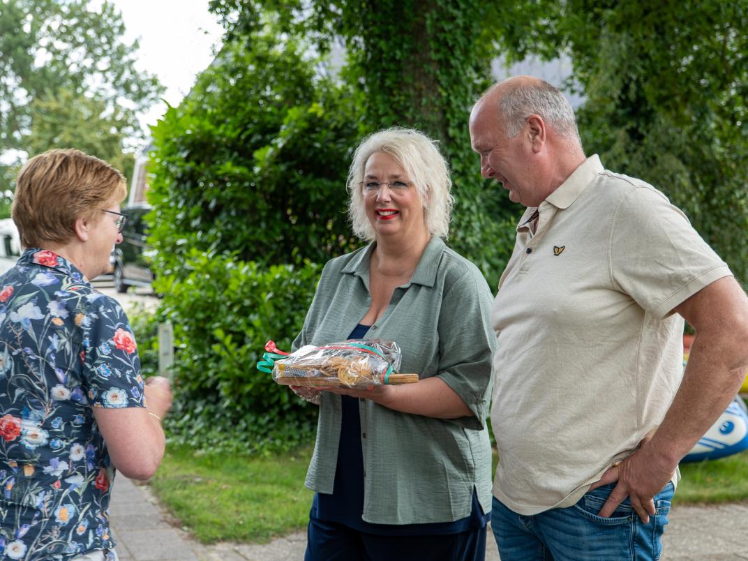 Drie personen staan buiten; links staat Maaike Prins (wethouder) en overhandigt iets aan een ander, met groen en een camper op de achtergrond.